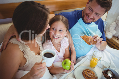 Parents sitting on bed with daughter and having breakfast