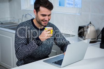 Man holding coffee mug while using laptop in the kitchen