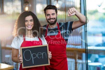 Smiling waitress and waiter standing with open sign board in cafe
