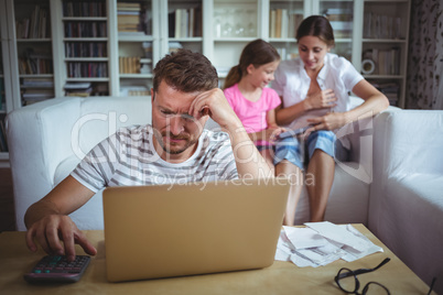 Worried man calculating bills while his wife and daughter sitting on sofa