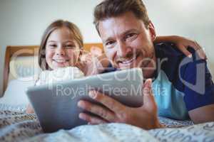 Father lying with daughter on bed and using digital tablet