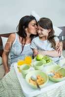 Mother kissing on daughter cheeks while having breakfast on bed