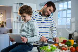 Father talking on mobile phone while chopping vegetables and son using digital tablet