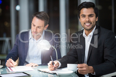 Businessman smiling at camera while colleague writing on paper