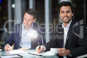 Businessman smiling at camera while colleague writing on paper