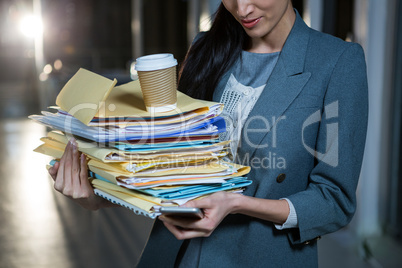 Businesswoman carrying stack of file folders while using mobile phone