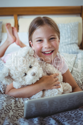 Happy girl lying on bed with a teddy bear and using digital tablet