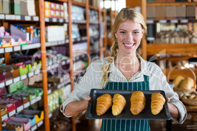 Smiling female staff holding tray of croissants in supermarket