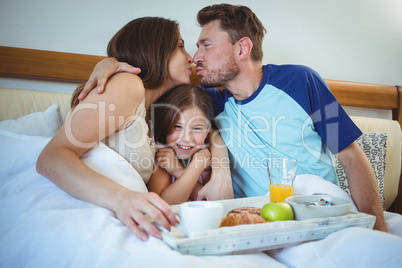 Parents kissing while sitting on bed with daughter and having breakfast