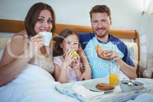 Parents sitting on bed with daughter and having breakfast