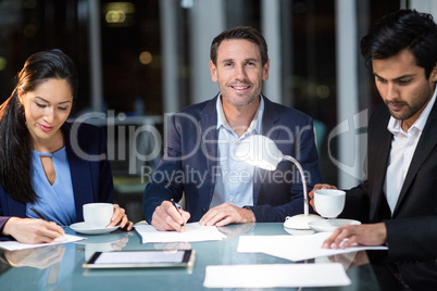 Businessman smiling at camera while colleagues writing on paper Businessman smiling at camera while colleagues writing on paper