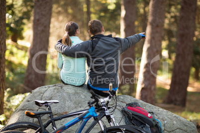 Biker couple sitting on rock pointing at distance in forest