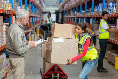 Warehouse manager noting on clipboard while female worker carrying cardboard boxes