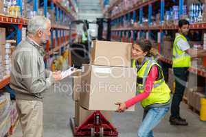 Warehouse manager noting on clipboard while female worker carrying cardboard boxes