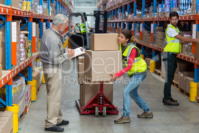 Warehouse manager noting on clipboard while female worker carrying cardboard boxes