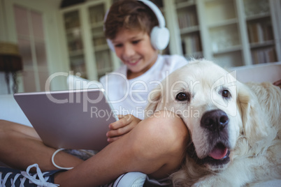 Boy sitting on sofa with pet dog and listening to music on digital tablet