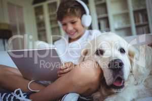 Boy sitting on sofa with pet dog and listening to music on digital tablet