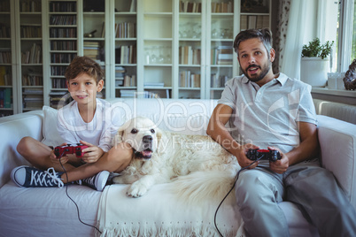 Father and son sitting on sofa with pet dog and playing video games