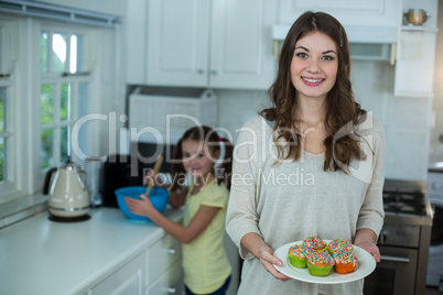 Woman holding a plate of cupcakes while her daughter preparing breakfast