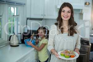 Woman holding a plate of cupcakes while her daughter preparing breakfast