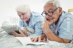 Senior couple reading a book and using digital tablet on bed