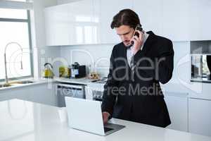 Businessman using laptop while talking on mobile phone in kitchen