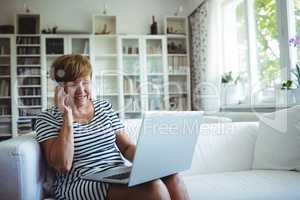 Senior woman talking on mobile phone while using laptop in living room