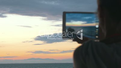 Woman takes photos of the beautiful scenery of the sea and evening sky with her tablet at sunset