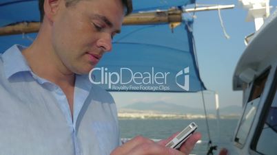 Man is typing on his smartphone while he sails on the boat in summer sunny day