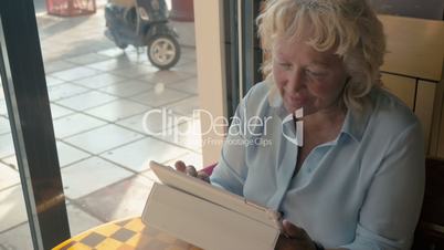 Mature woman sitting in a cafe and enjoys her tablet., outside the window the summer, sunny day