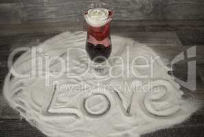 Glas mit Rose und Love Schriftzug im Sand