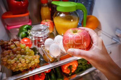 Woman takes the apple from the open refrigerator.