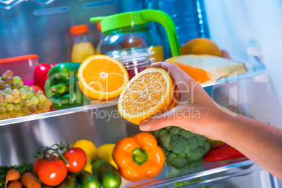 Woman takes the orange from the open refrigerator.