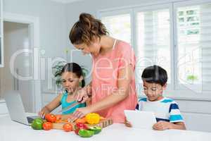 Kids using digital tablet and laptop while mother chopping vegetables in kitchen