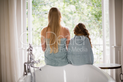 Rear view of mother and daughter sitting on bathtub