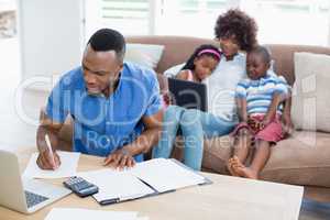 Worried man calculating bills while his wife and kids using digital tablet on sofa