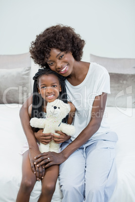 Smiling mother and daughter sitting with arm around on bed at home