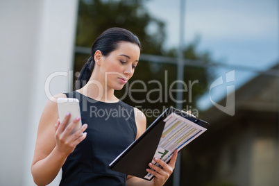 Businesswoman holding mobile phone and looking at clipboard