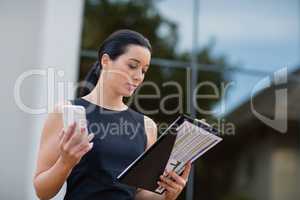 Businesswoman holding mobile phone and looking at clipboard
