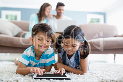 Siblings using digital tablet while lying on rug in living room