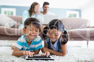 Siblings using digital tablet while lying on rug in living room