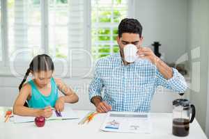Daughter doing her homework and father having a cup of black tea while reading newspaper
