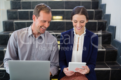 Businessman and woman sitting on steps using laptop and digital tablet