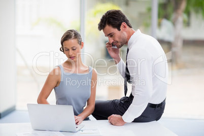 Businesswoman using laptop while colleague talking on mobile phone
