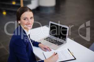 Businesswoman sitting at desk with clipboard and laptop