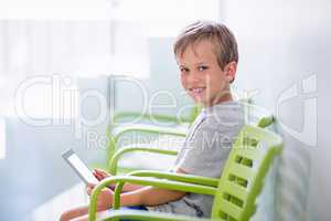 Portrait of boy sitting on chair using digital tablet in corridor