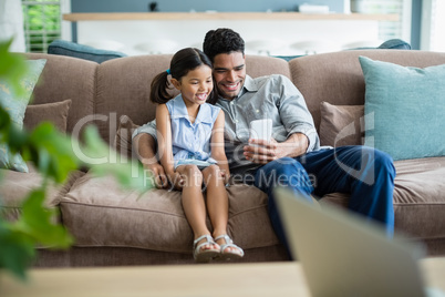 Father and daughter sitting on sofa and using mobile phone