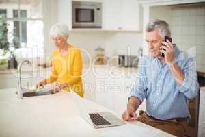 Senior man talking on mobile phone while woman washing plate in kitchen