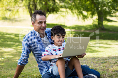 Boy sitting on his fathers lap and using laptop in park