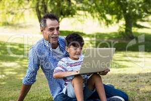 Boy sitting on his fathers lap and using laptop in park